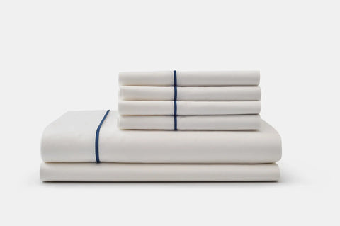 The Cotton Piping Design Bed Sheet Set by American Blossom Linens features luxurious white cotton sheets and pillowcases with a thin dark blue stripe, neatly stacked against a plain white background.