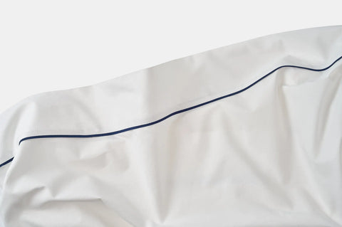 A close-up of the Cotton Piping Design Bed Sheet Set by American Blossom Linens, made from USA-grown cotton, featuring a single thin dark blue line across smooth white fabric with soft folds and gentle wrinkles.
