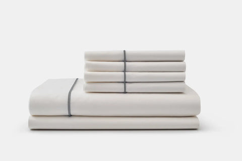 The American Blossom Linens Cotton Piping Design Bed Sheet Set, featuring luxurious white cotton sheets and pillowcases with a thin dark gray trim, is neatly folded and stacked on a plain white background.