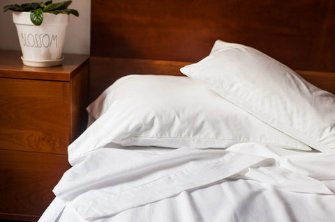 A neatly made bed features the Cotton Single Fitted Bed Sheet by American Blossom Linens, crafted from luxurious USA-grown cotton, topped with two white pillows. A wooden nightstand holds a potted plant labeled BLOSSOM by the wooden headboard.