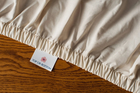 A close-up of the American Blossom Linens Cotton Single [Separate] Fitted Bed Sheet, made from USA-grown cotton, featuring a TOP OR BOTTOM tag beside a small pink flower on a wooden surface.