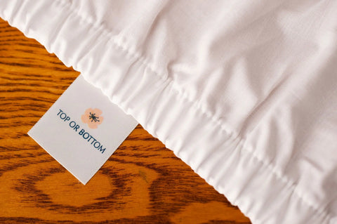 A close-up of American Blossom Linens’ Cotton Single [Separate] Fitted Bed Sheet, made from USA-grown cotton, showing a “TOP OR BOTTOM” tag with a flower illustration on a wooden surface.