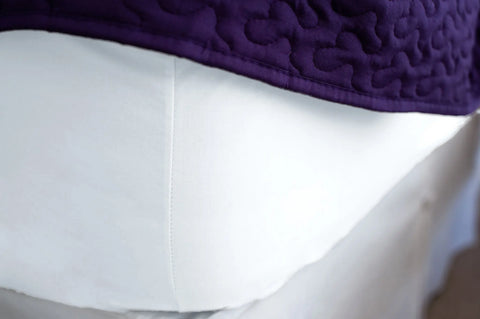 Close-up of a neatly made bed featuring the Cotton Single Fitted Bed Sheet by American Blossom Linens, crafted from luxurious USA-grown cotton and topped with a textured purple quilt.