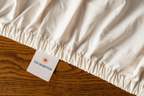 A close-up of the cream-colored Cotton Single Fitted Bed Sheet by American Blossom Linens, made from premium USA grown cotton, with elastic edges and a “TOP OR BOTTOM” tag featuring a small floral design on a wooden surface.
