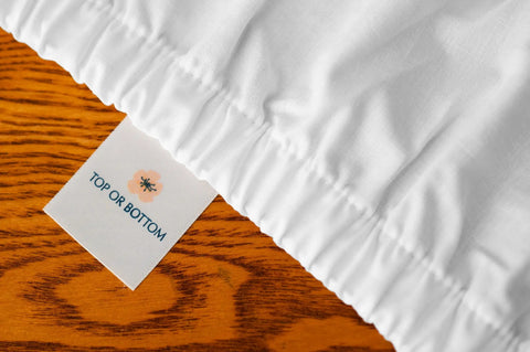 A close-up of the Cotton Single [Separate] Fitted Bed Sheet by American Blossom Linens, in white, on a wooden surface—showing a fabric tag with a flower and “TOP OR BOTTOM.”—crafted from premium USA grown cotton.