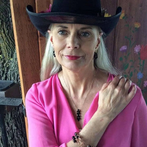 A woman with long gray hair in a black cowboy hat and bright pink blouse wears an Alexa Martha Designs Gold Wire Bead-Woven Rock Candy Crystal Bar Necklace, resting her hand on her shoulder near a tree and painted wooden wall.