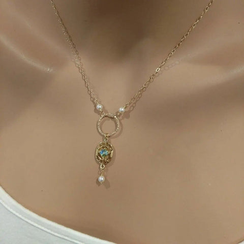 The 14K Gold-Filled Hammered Open Circle Filigree Necklace by Alexa Martha Designs features Swarovski crystal pearls and an iridescent stone, elegantly displayed on a beige mannequin in a white top. Each piece is handmade in Montana.