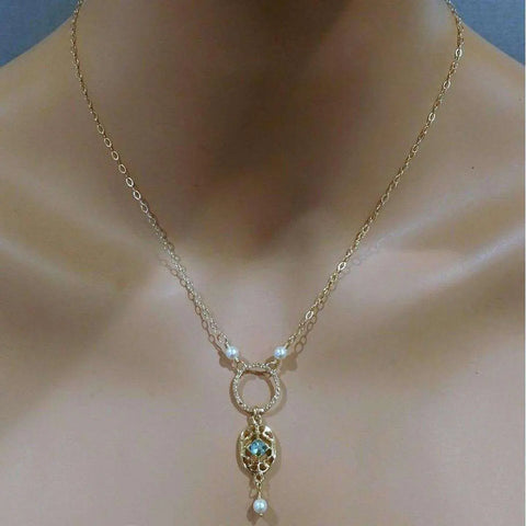 The 14K Gold-Filled Hammered Open Circle Filigree Necklace by Alexa Martha Designs features Swarovski crystal pearls and a blue gemstone pendant with a dangling pearl, all handmade in Montana and shown on a mannequin’s neck.