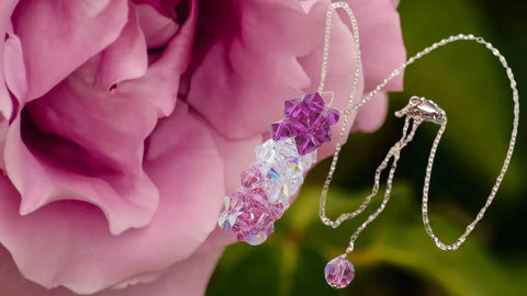 The 14 K Rose Gold Fill Super Sparkly Mom Crystal Pillar Pendant Necklace by Alexa Martha Designs is displayed in front of a blooming pink rose against a soft green background.