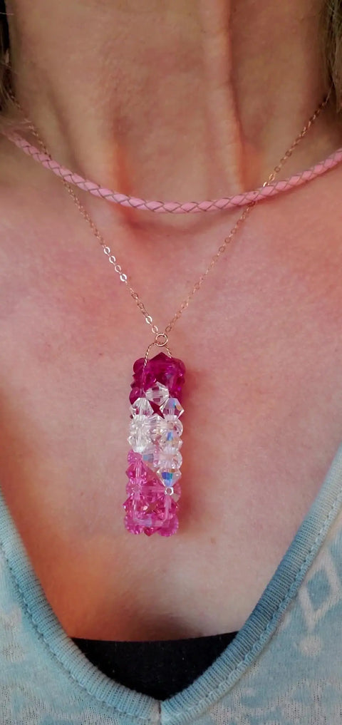 A close-up of a neck adorned with a pink braided choker and Alexa Martha Designs 14K Rose Gold Fill Super Sparkly Mom Crystal Pillar Pendant Necklace, layered over light blue clothing.