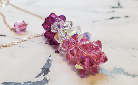 A close-up shows the 14 K Rose Gold Fill Super Sparkly Mom Crystal Pillar Pendant Necklace by Alexa Martha Designs, featuring faceted crystals in pink, purple, and clear gradients on a marble-inspired surface with silver streaks.