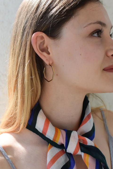 A woman with straight, light brown hair wears Sarah Cecelias GOLD GEO HOOPS | LARGE—14k gold-plated geometric hoop earrings—and a colorful striped neck scarf. Facing right, sunlight highlights her face and hair.