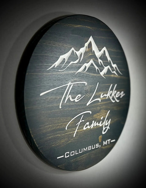 The B. James Design & Woodwork 15 Round Custom Family or Name Sign features engraved white text reading The Lukkes Family, Columbus, MT below a mountain design—an ideal personalized décor piece for your home.
