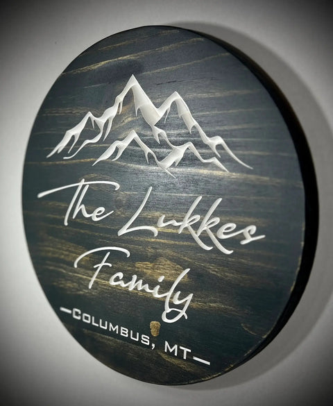 The 15 Round Custom Family or Name Sign by B. James Design & Woodwork features engraved snowy mountain peaks above your family name and location in white script and block lettering on dark rustic wood—perfect personalized décor.
