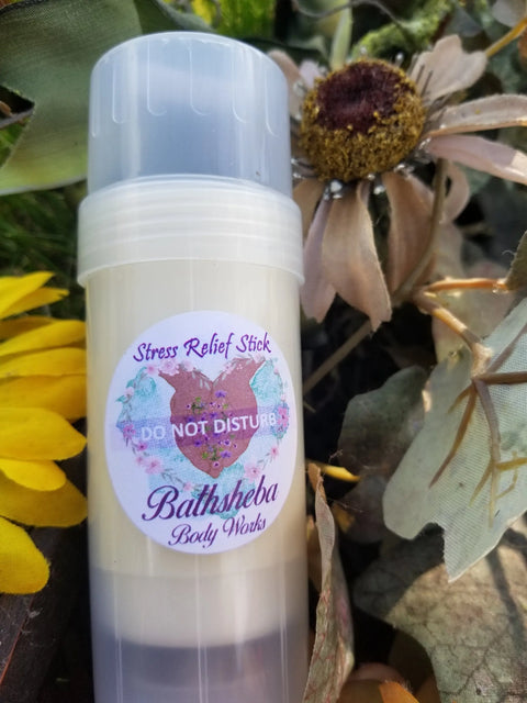 A tube labeled Moisture Balm - Do Not Disturb (Relief Stick) from Bathsheba Body Works sits among yellow petals and dried flowers. The label features a blue heart with purple accents, highlighting this soothing essential oil remedy.