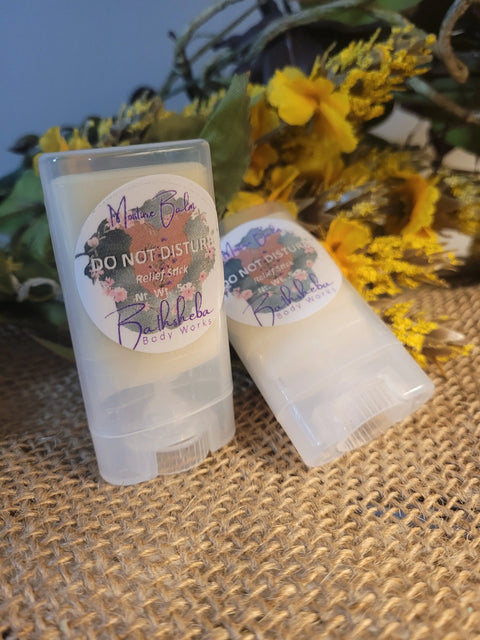 Two translucent mini tubes labeled Moisture Balm - Do Not Disturb (Relief Stick) from Bathsheba Body Works rest on burlap amid yellow artificial flowers and greenery—a soothing essential oil remedy for stress relief.