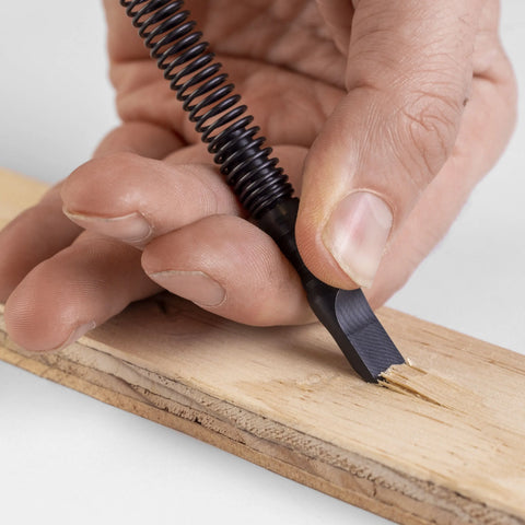 A close-up of a hand using the Hammerless Impact Technology Wood Chisel with a metal blade and coiled handle to carve a groove into a light wooden board. This American-made chisel features a hammerless design for easy, precise work.