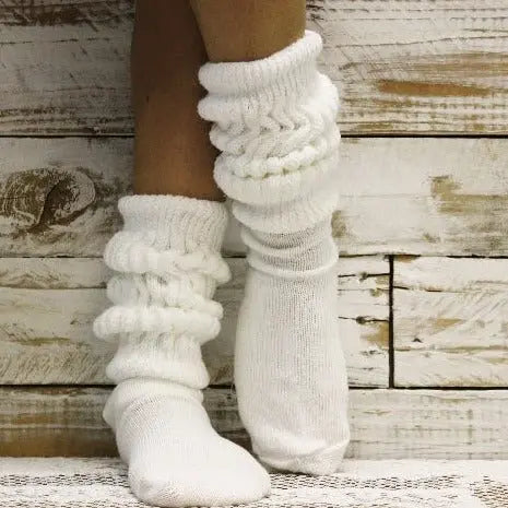 A person wears Catherine Coles 3 Pairs Bundle ULTIMATE Slouch Socks in white, standing with legs crossed before a rustic wooden wall and capturing classic 80s fashion vibes.