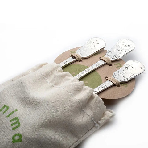 Three Arctic Friends baby spoons by Kleynimals, featuring engraved animal faces, peek out of a beige pouch with green text. The 3-piece stainless steel flatware set is secured with rubber bands and displayed on a round cardboard backing.