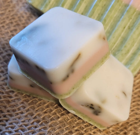 Three square, layered all natural soap bars from Bathsheba Body Works are stacked on a brown surface. In the background, the All Natural Soap Bar - Herbal Caress in striped green and white adds a touch of herbal freshness.
