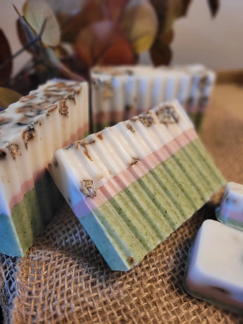 Three handmade Bathsheba Body Works All Natural Soap Bar - Herbal Caress, with green, pink, and white stripes, sit on burlap and are accented by dried flowers with blurred brown leaves in the background.