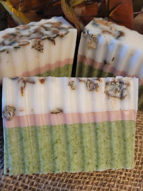A close-up of Bathsheba Body Works All Natural Soap Bar - Herbal Caress showcases handmade bars with green, pink, and white layers, textured edges, and dried herbs on top, elegantly displayed on burlap with autumn leaves behind.