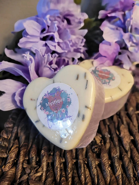 Two heart-shaped Moisture Bar - Mystique soaps by Bathsheba Body Works rest on a woven surface, surrounded by light purple artificial flowers. Enriched with Abyssinian Oil, each bar contains visible lavender pieces and a decorative top sticker.