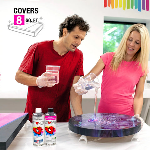 A man and woman in pink shirts and gloves pour ArtResin’s 32 Oz Epoxy Resin Starter Pack onto a circular artwork. Two ArtResin bottles sit on the table beneath a rainbow wall graphic that reads “COVERS 8 SQ. FT.”—ideal for resin crafting.