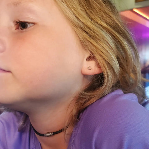 A child with light brown hair wears a heart-shaped earring, a purple shirt, and a black 3mm Bolo Leather Snap Off Choker by Alexa Martha Designs. The softly lit background features warm and purple tones.