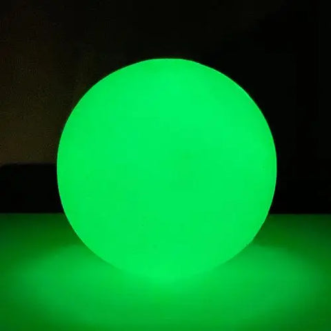 The SodaPup 4 PUP-X EDispenser Glow Ball Treat Dispenser glows green in the dark, illuminating a soft light around its base as it sits on a flat surface.
