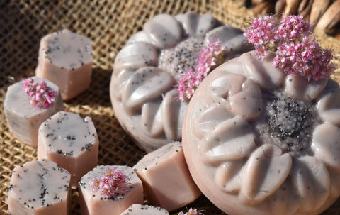Handmade round and hexagonal Body Butter Scrub - Sunny Days bars by Bathsheba Body Works, pink with black specks and some topped with pink flower clusters, displayed on burlap—a perfect touch for your natural skincare routine.