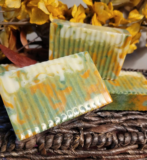 Three rectangular bars of Bathsheba Body Works All Natural Soap - Ginger Turmeric (Herbal Soap), featuring green, orange, and white swirls, rest on a woven brown surface with yellow dried flowers in the background.