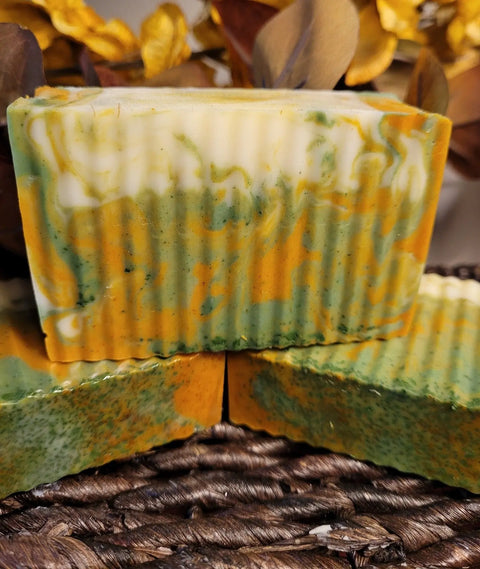 Two handmade bars of Bathsheba Body Works All Natural Soap - Ginger Turmeric feature green, orange, and cream marbling with wavy edges. Set on dried leaves and a woven surface, these herbal soaps showcase true artisanal appeal.