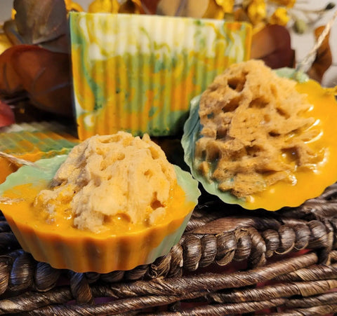 Two All Natural Soap - Ginger Turmeric bars from Bathsheba Body Works rest on a woven surface: a rectangular, marbled green-yellow-orange bar, and round soaps with rough tan tops and orange bases in green cupcake liners.