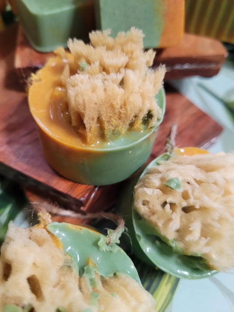 Round bars of Bathsheba Body Works’ All Natural Soap – Ginger Turmeric feature natural sponges embedded on top, contrasting beautifully with the smooth, colorful herbal soap, and are displayed on a wooden tray.