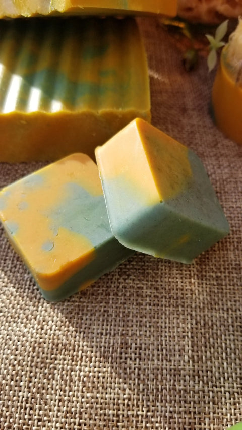 Two square bars of Bathsheba Body Works All Natural Soap - Ginger Turmeric, featuring orange and green marbling, rest on burlap; a larger ridged bar and a slice of orange appear in the background.