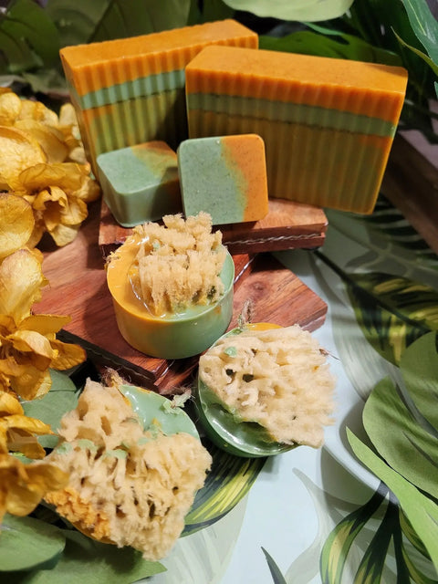 Bathsheba Body Works’ All Natural Soap - Ginger Turmeric (Herbal Soap) in green, orange, and yellow hues is displayed on wooden boards with small soaps topped by sponges, accented with yellow dried flowers and green leaves for a botanical vibe.