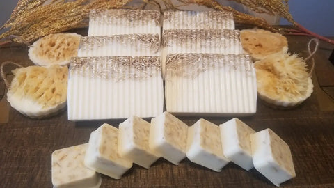 An assortment of Bathsheba Body Works’ All Natural Soap Bar - Country Life rests on wood, with rectangular exfoliating bars, round loofah soaps, and small squares—each highlighting natural botanicals within.