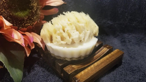 The All Natural Soap Bar - Country Life by Bathsheba Body Works rests on a wooden soap dish with a sea sponge on top, paired with a peach flower for exfoliation, set against dark fabric.