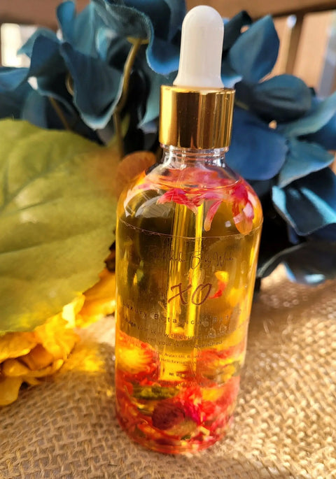 A clear glass dropper bottle of Bathsheba Body Works Body Oil - XO, filled with golden oil and red flower petals, rests on textured burlap with large blue and yellow artificial flowers in the background.