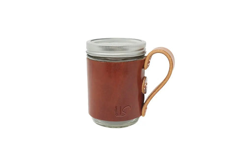 The Urban Southern Mason Jar Mug features a clear body, metal lid, and brown leather sleeve with a right-hand handle and subtle embossed logo for a vintage look.
