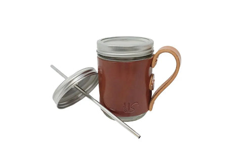 The Urban Southern Mason Jar Mug features a brown leather sleeve with handle, metal lid, and straw. The leather-wrapped mug is shown with the lid resting against it, all set on a plain white background.