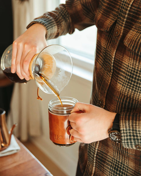 A person in a plaid shirt pours coffee from a glass carafe into an Urban Southern Mason Jar Mug while standing near a window.