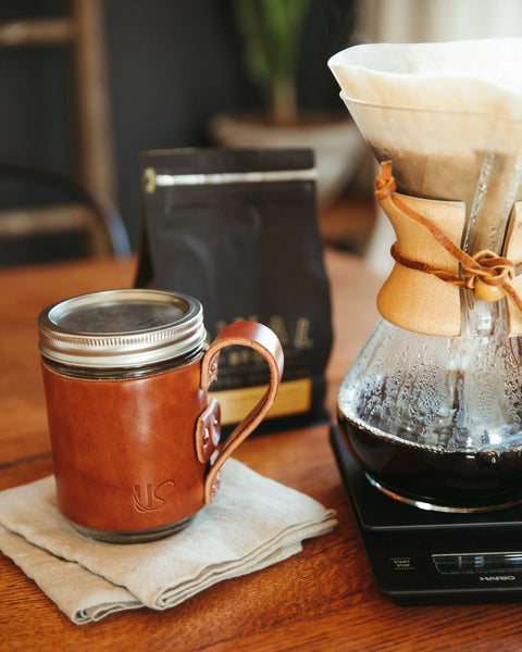 An Urban Southern Mason Jar Mug sits on a folded napkin next to a Chemex coffee maker on a digital scale, with a bag of coffee beans in the background atop a wooden table.