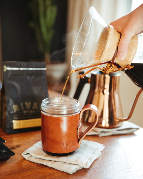 A hand pours hot coffee from a Chemex into an Urban Southern Mason Jar Mug on a wooden table, with a bag of Rival Bros. coffee and a copper kettle in the background. Steam rises from the mug.