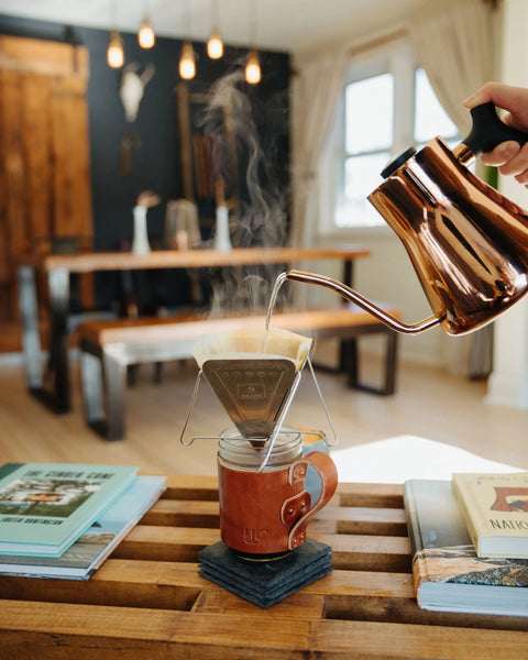 A hand pours hot water from a copper kettle over coffee grounds in a pour-over setup, with steam rising above an Urban Southern Mason Jar Mug on a wooden table in a cozy, sunlit living space surrounded by books and coasters.