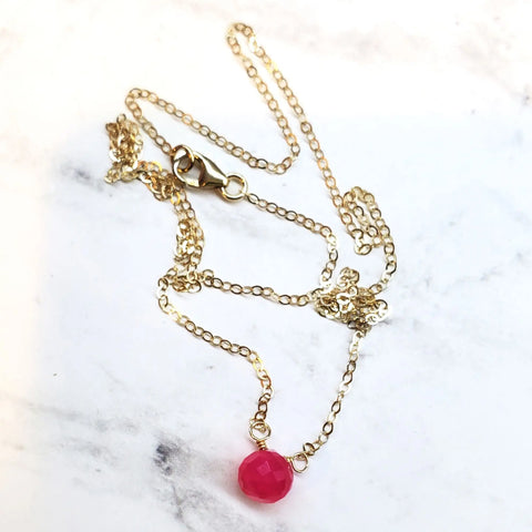 The Tiny Hot Pink Chalcedony Gold Filled Necklace by Alexa Martha Designs features a single faceted pink bead pendant and a small lobster clasp, handcrafted for elegance and displayed on white marble.