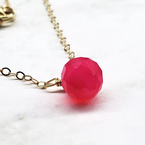 The Tiny Hot Pink Chalcedony Gold Filled Necklace by Alexa Martha Designs features a faceted round chalcedony pendant on a delicate gold filled chain, shown on a white marble surface.
