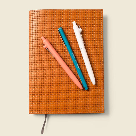 The CASUPO Refillable A5 Leather Notebook Cover – Tan Limited Edition lies closed with three pens—coral, teal, and white—placed diagonally on top and a gray ribbon bookmark, set against a light beige background.