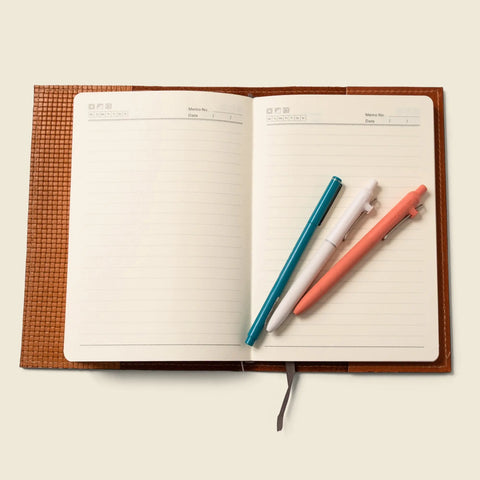 The CASUPO Refillable A5 Leather Notebook Cover – Tan Limited Edition lies open on a light beige surface, showing lined pages with a blue, white, and pink pen placed diagonally across them.